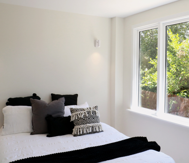 Newly renovated bedroom with bed. Bed has white bed linen, black white and grey cushions and large window overlooking a garden.